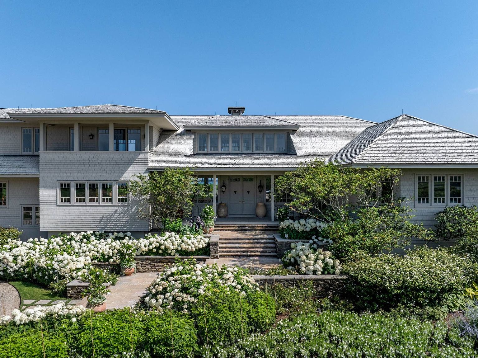 This elegant shingle-style residence features a symmetrical facade with a prominent central entryway and a wide, welcoming front porch. The home is surrounded by lush, tiered landscaping filled with abundant white hydrangea bushes and stone retaining walls that lead up to the front doors. The perspective is captured from a slightly elevated angle, showcasing the expansive roofline and the harmonious integration of the architecture with the manicured garden.