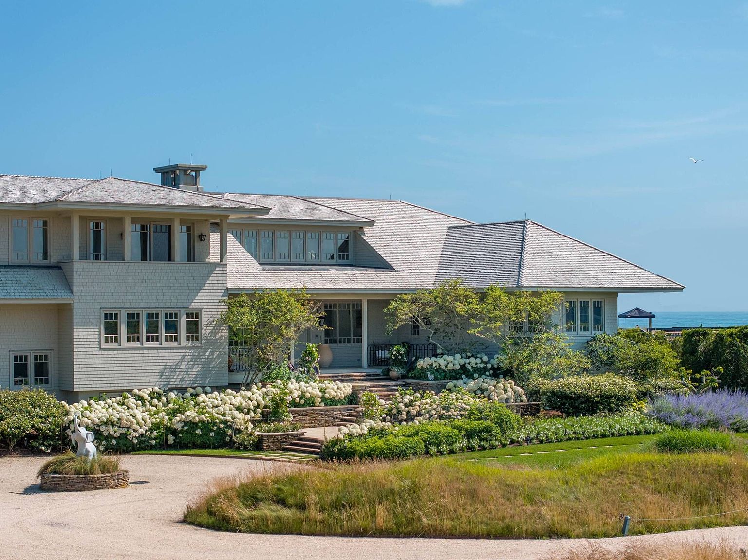 This expansive coastal-style residence features light-colored shingle siding and a multi-gabled roofline, set against a clear blue sky. The front of the home is framed by lush landscaping, including abundant white hydrangea bushes and stone steps leading to the main entrance. A gravel driveway curves through the foreground, complemented by a small decorative sculpture and a view of the ocean in the distance.
