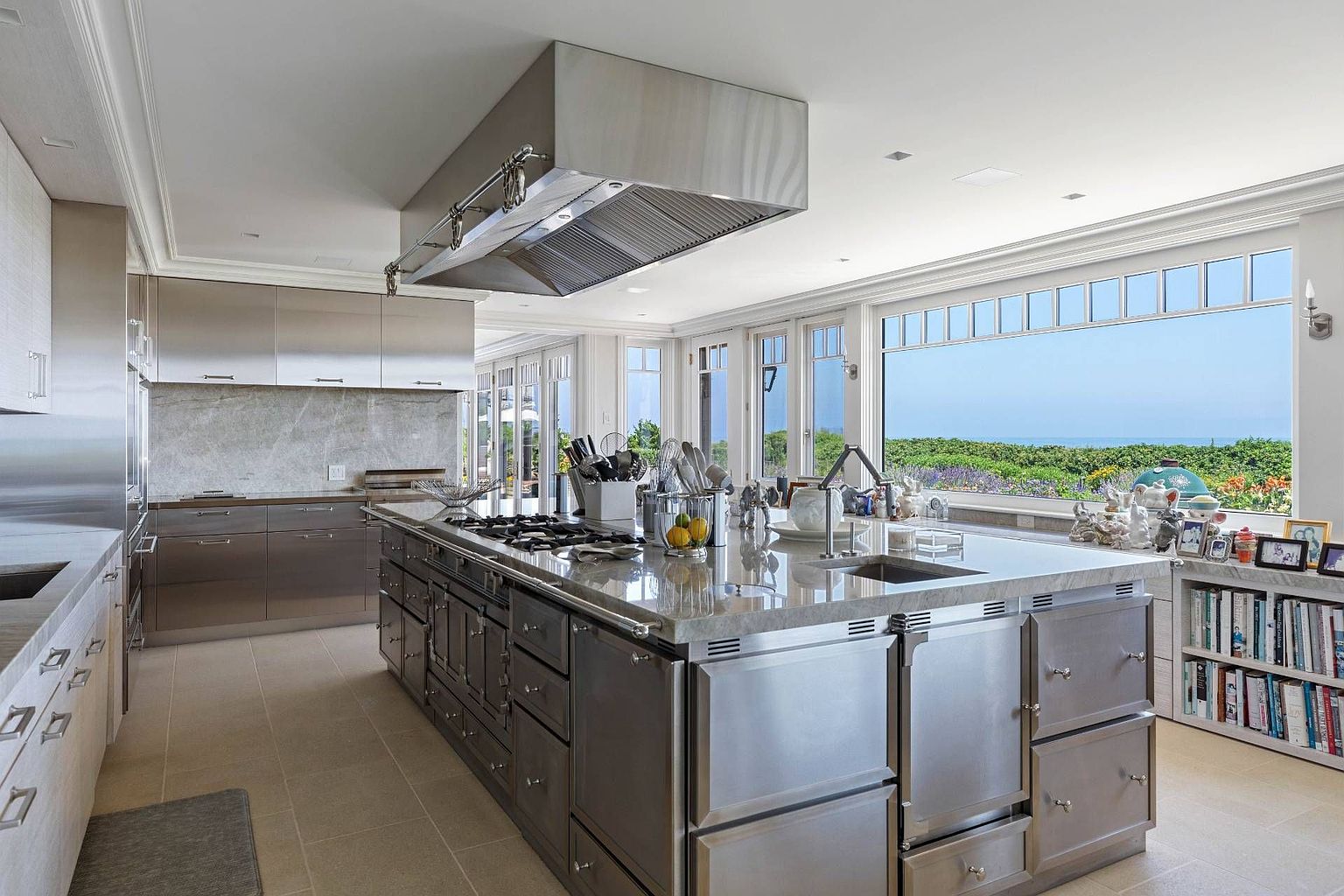 This professional-grade kitchen features an expansive stainless steel island equipped with a built-in gas cooktop and a large overhead ventilation hood. The space is defined by its sleek metallic cabinetry, stone countertops, and a wall of large windows that provide an unobstructed view of the outdoor landscape and horizon. A built-in bookshelf at the end of the island adds a functional touch, while the neutral tile flooring and clean lines create a sophisticated, industrial-inspired aesthetic.