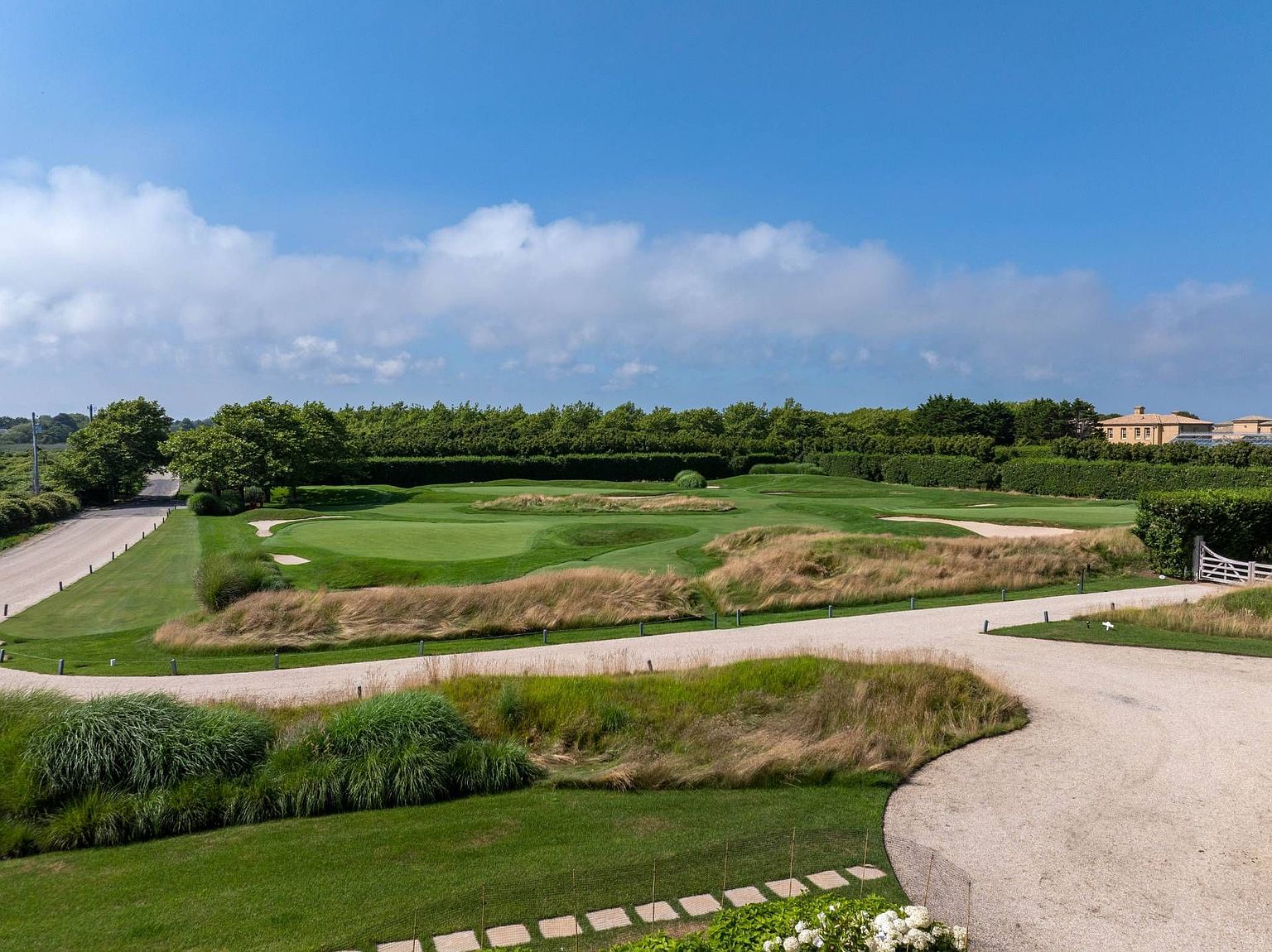 This elevated perspective captures a sprawling, meticulously manicured golf course landscape characterized by rolling green fairways, sand bunkers, and patches of tall, ornamental grasses. A gravel path winds through the foreground, leading the eye toward a dense, mature hedge line that frames the property. The scene is bathed in bright, natural daylight under a clear blue sky, evoking a sense of expansive, serene luxury.