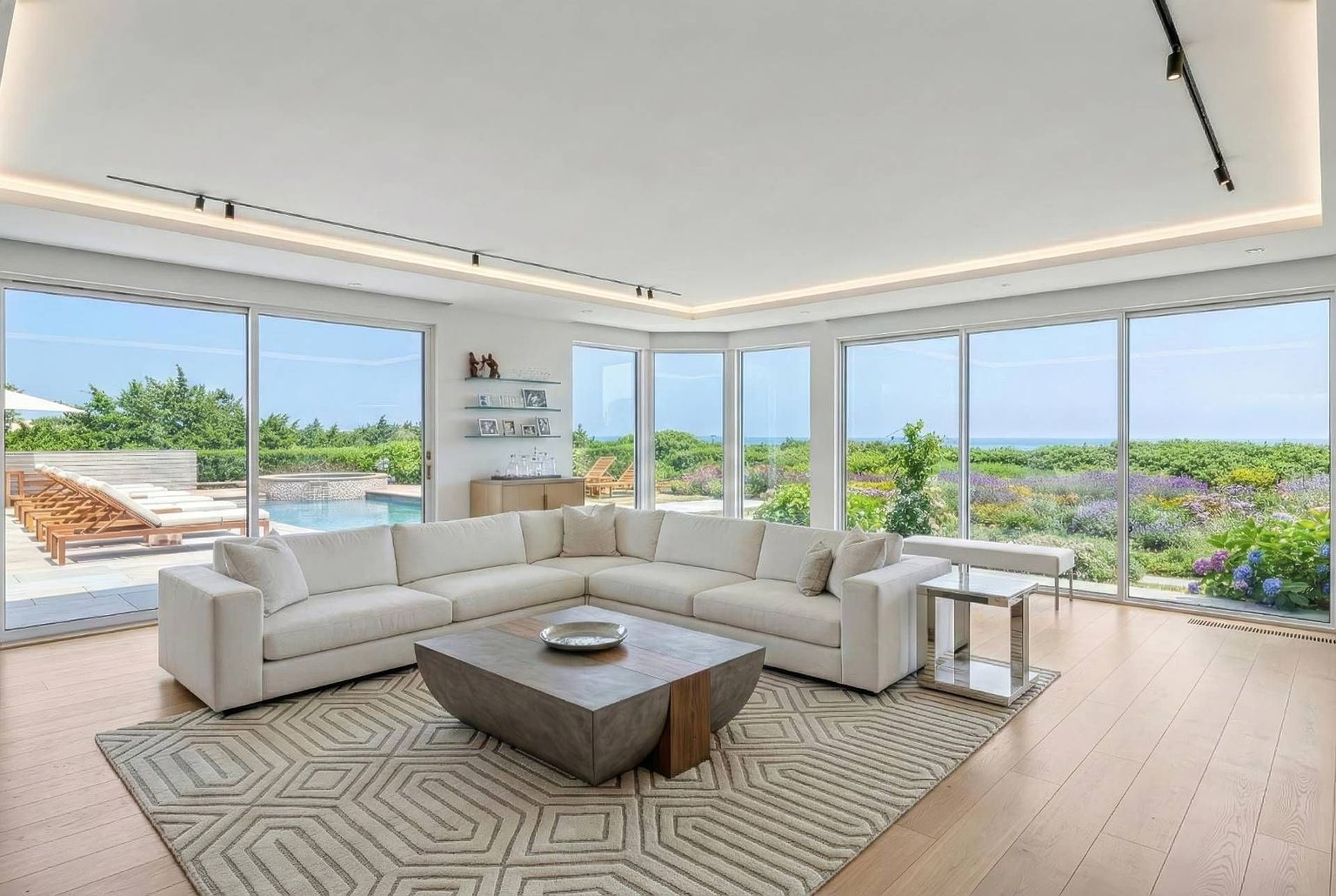 This bright and airy living room features a large, neutral-toned sectional sofa arranged around a substantial, low-profile coffee table with a geometric-patterned area rug. The space is defined by expansive, floor-to-ceiling glass walls that provide panoramic views of the outdoor pool area and the lush, blooming landscape beyond. The minimalist aesthetic is complemented by light wood flooring, recessed lighting, and a clean, open-concept layout that seamlessly connects the interior to the exterior environment.