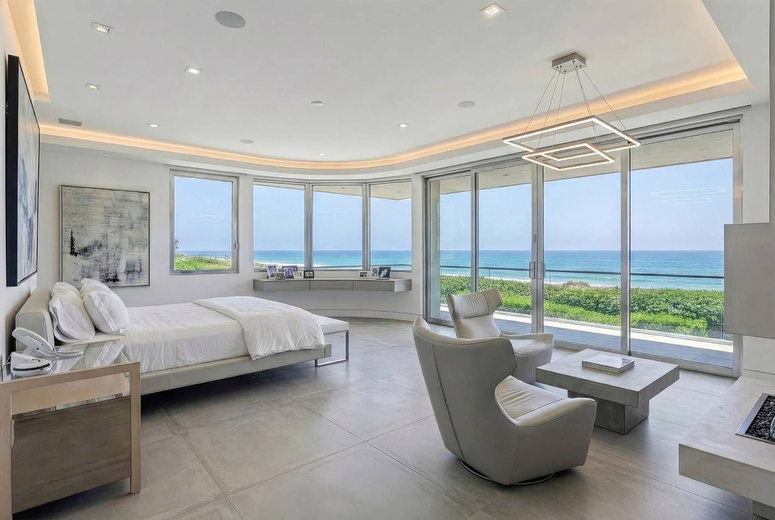 This expansive primary bedroom features a contemporary design with large windows that provide panoramic views of the ocean. The room is furnished with a low-profile bed, two modern leather armchairs, and a minimalist stone coffee table, all set against a neutral color palette. A sleek, modern chandelier hangs from the recessed ceiling, while a floating shelf along the curved wall adds a functional and aesthetic touch to the space.