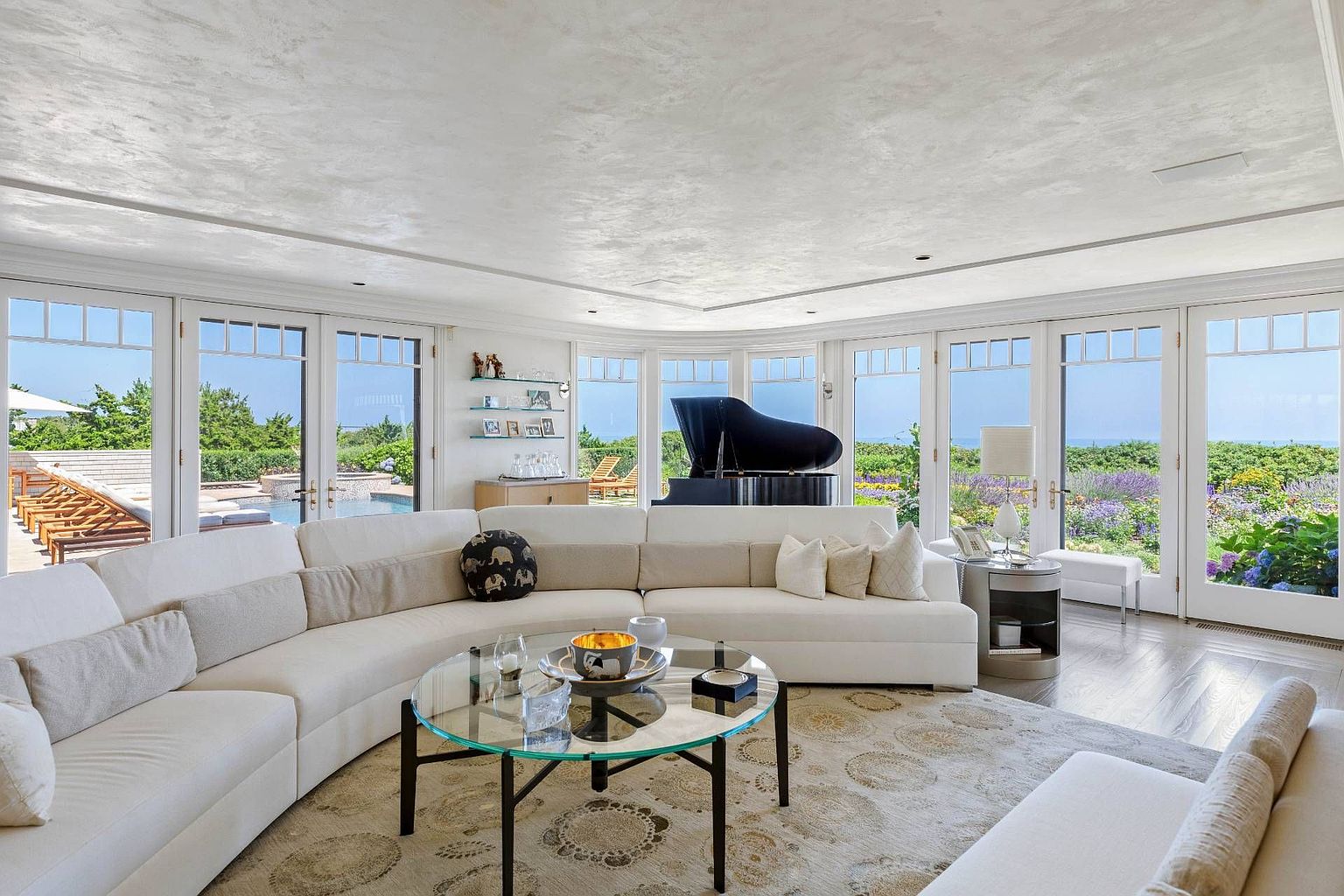 This bright and airy living room features a large, curved white sectional sofa arranged around a circular glass-topped coffee table. The space is defined by a series of tall windows and glass doors that provide panoramic views of the lush garden and pool area outside. A grand piano sits prominently near the windows, while a patterned area rug adds texture to the light-toned wood flooring, creating an elegant and inviting atmosphere.