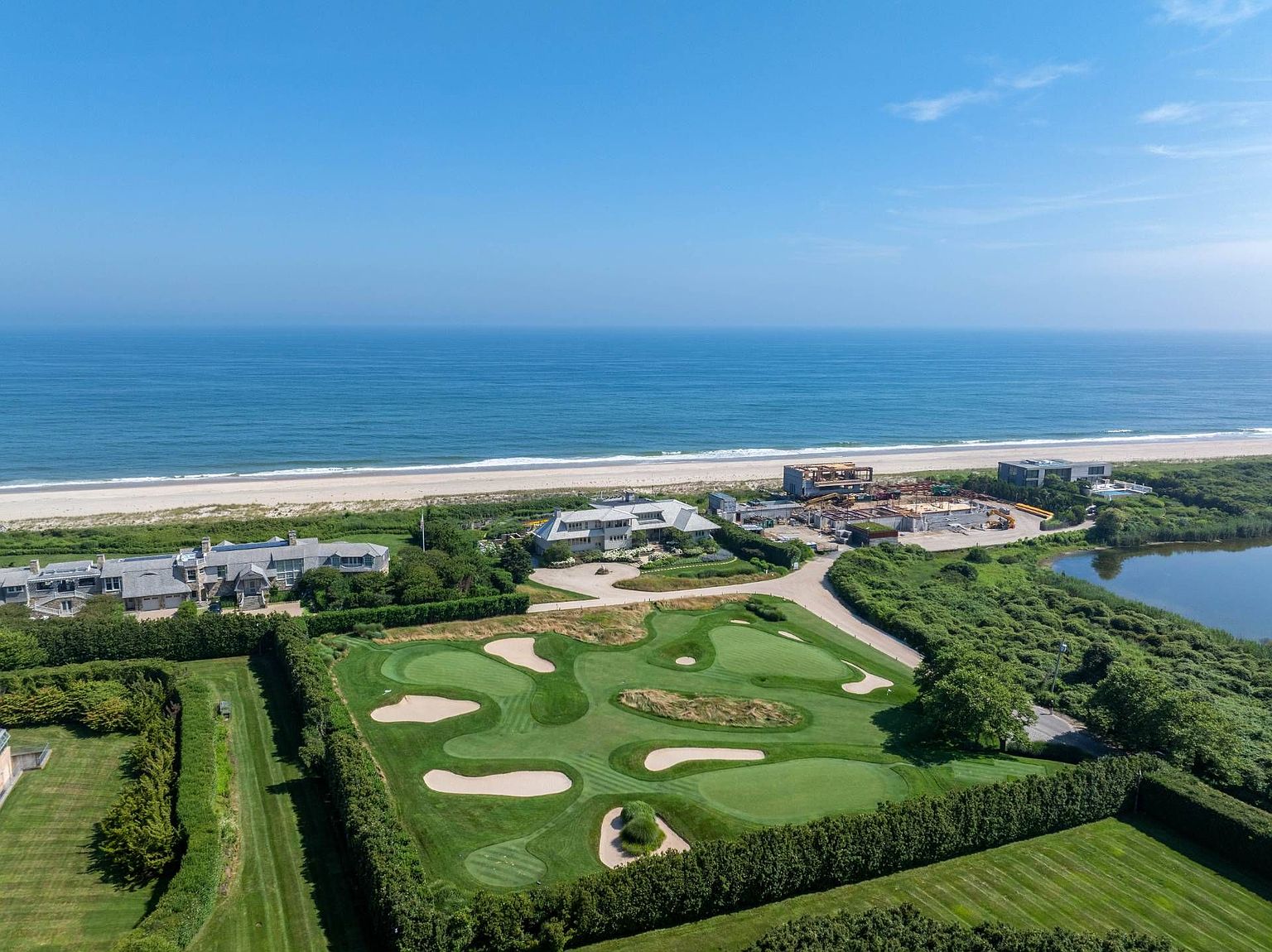 This expansive aerial view captures a luxurious coastal estate featuring a private golf practice green, multiple large residences, and direct access to a pristine sandy beach. The property is surrounded by lush greenery and mature hedges, with a construction site visible nearby, highlighting the ongoing development of this exclusive waterfront location. The cinematic perspective emphasizes the vastness of the ocean horizon and the seamless integration of high-end landscaping with the natural coastal environment.