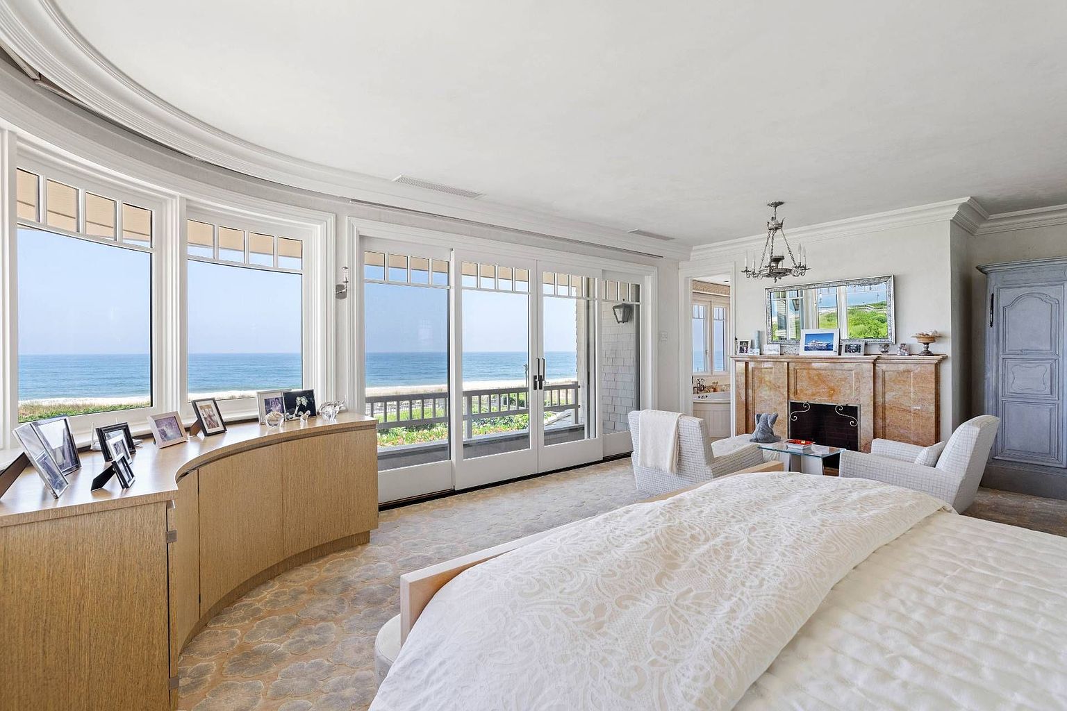This spacious primary bedroom features a curved light wood console table and a large bed with a textured white coverlet. A prominent stone fireplace with a decorative mantel serves as a focal point, while expansive windows and glass doors provide panoramic views of the ocean and beach. The room is decorated in a neutral, elegant palette, creating a serene and luxurious atmosphere.