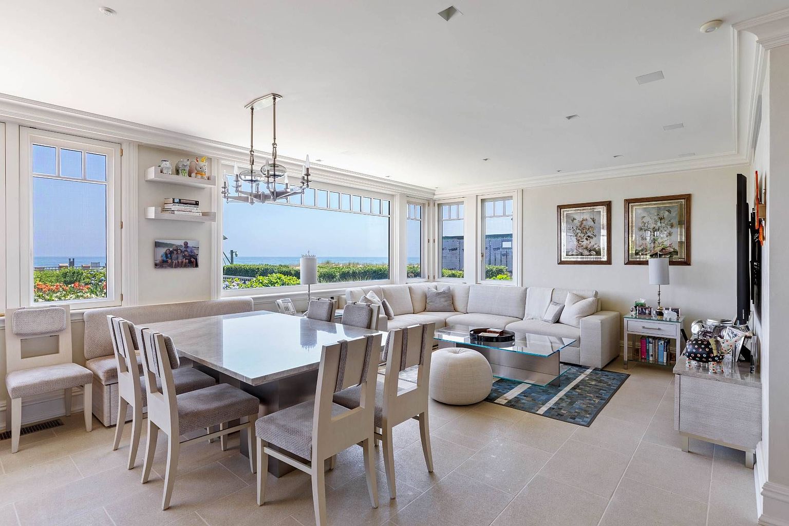 This open-concept living and dining area features a bright, airy aesthetic with neutral tones and large windows that frame a scenic ocean view. The space includes a stone-topped dining table with matching chairs, a plush sectional sofa, and a glass coffee table, all arranged on light-colored tile flooring. A modern chandelier hangs above the dining area, while the room is accented by framed artwork and decorative shelving, creating a sophisticated and inviting coastal atmosphere.