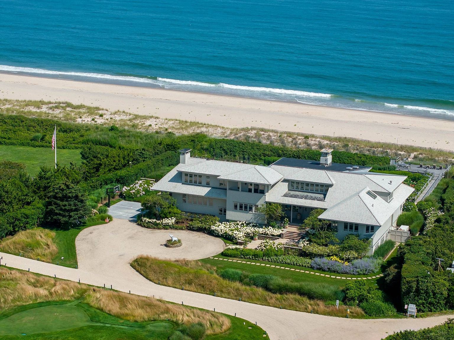 This high-angle aerial view captures a sprawling coastal estate nestled directly against a sandy beach and the ocean. The shingled residence features a multi-gabled roofline, a circular gravel driveway with a central sculpture, and lush, manicured landscaping with abundant white flowering shrubs. A private wooden walkway leads from the property toward the dunes, emphasizing the home's prime waterfront location and serene, secluded atmosphere.