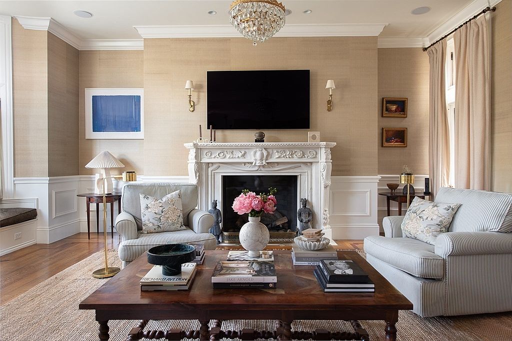 This elegant living room features a central fireplace with an ornate, carved mantel topped by a wall-mounted television. The space is anchored by a large, dark wood coffee table and two striped armchairs, all set upon a textured natural fiber rug. The walls are finished in a neutral, textured wallpaper, complemented by white wainscoting and a crystal chandelier that adds a touch of sophistication to the room.