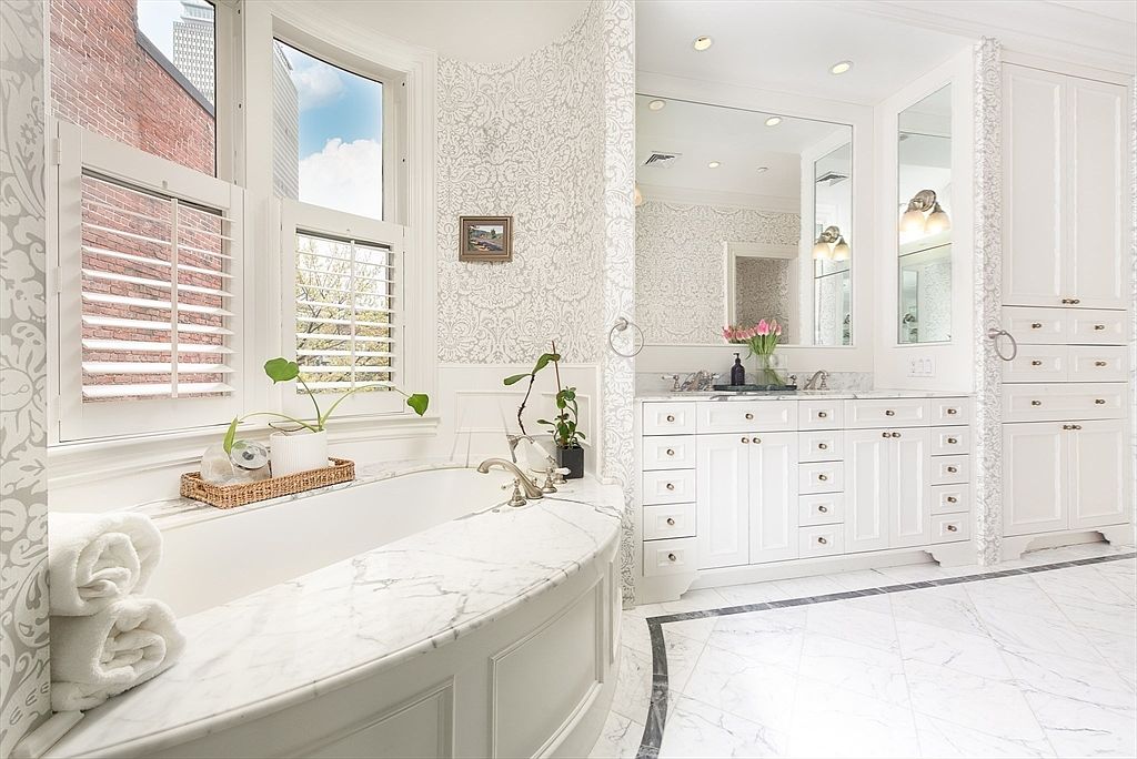 This elegant primary bathroom features a large, curved soaking tub with a stone surround positioned beneath tall windows with plantation shutters. The space is adorned with patterned wallpaper, a double vanity with stone countertops, and a large mirror that reflects the bright, airy atmosphere. The floor is finished with light-colored tile featuring a dark decorative border, creating a sophisticated and luxurious aesthetic.