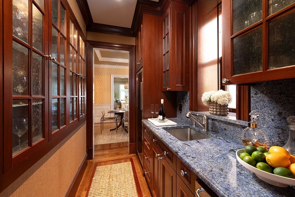 This elegant butler's pantry features rich, dark-toned cabinetry with glass-fronted upper doors and a striking blue-toned stone countertop. A stainless steel sink is integrated into the counter, which is accented by a bowl of fresh citrus and a decorative vase of white flowers. The space leads through a doorway into a formal dining area, showcasing a cohesive and sophisticated design aesthetic.