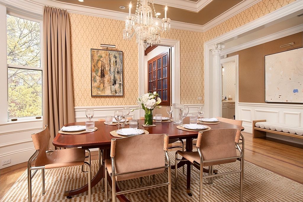 This elegant dining room features a rich wood table surrounded by modern chairs with leather-like upholstery and metal frames. The space is adorned with patterned wallpaper, a crystal chandelier, and a large abstract painting, all set against a backdrop of white wainscoting. A large window with neutral curtains allows natural light to fill the room, creating a sophisticated and inviting atmosphere.