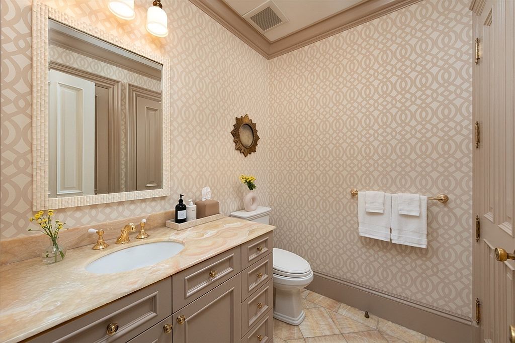 This elegant powder room features patterned wallpaper, a stone-topped vanity with a single undermount sink, and a classic toilet. The space is accented by a decorative mirror, a small vase with flowers, and a wall-mounted towel bar holding white linens. The perspective is a straight-on view of the vanity and toilet area, highlighting the sophisticated, neutral color palette and refined finishes.