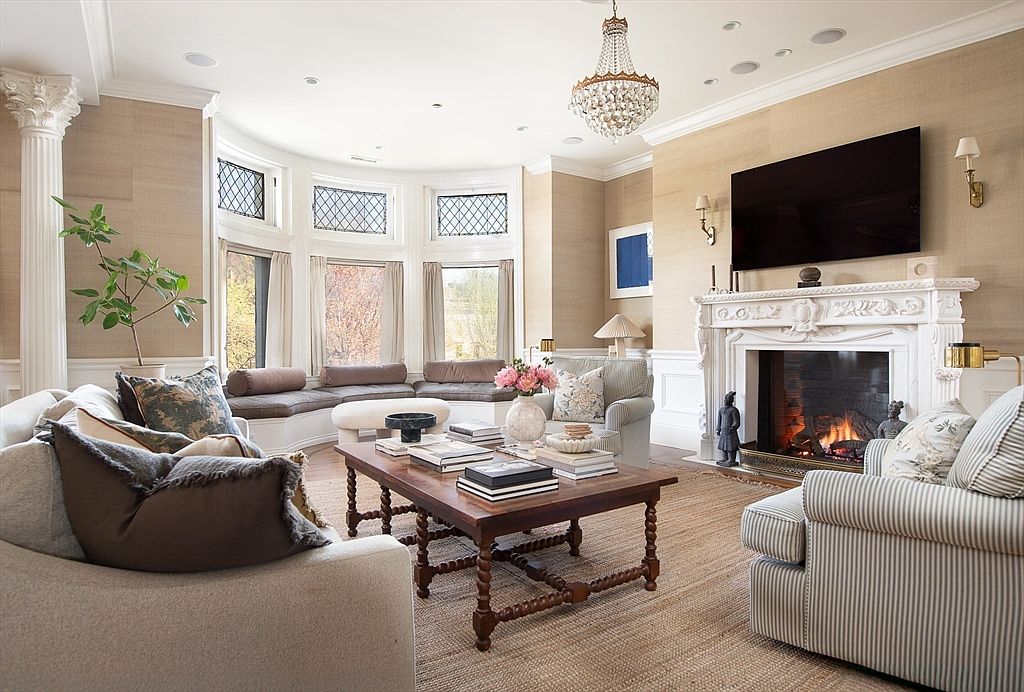 This elegant living room features a sophisticated neutral color palette with textured wall coverings and a prominent fireplace with an ornate white mantel. A large bay window area with built-in bench seating provides ample natural light, while a crystal chandelier serves as a focal point above the central seating arrangement. The room is furnished with a mix of plush sofas and a wooden coffee table, creating a warm and inviting atmosphere suitable for formal entertaining.