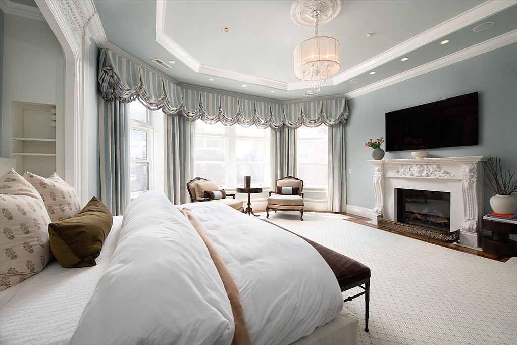 This elegant primary bedroom features a tray ceiling with a central chandelier and soft blue wall tones. A large, ornate fireplace with a television mounted above serves as a focal point, while a bay window area with custom striped valances provides a cozy seating nook. The room is furnished with a plush bed, a dark bench at the foot, and two armchairs, creating a sophisticated and tranquil retreat.