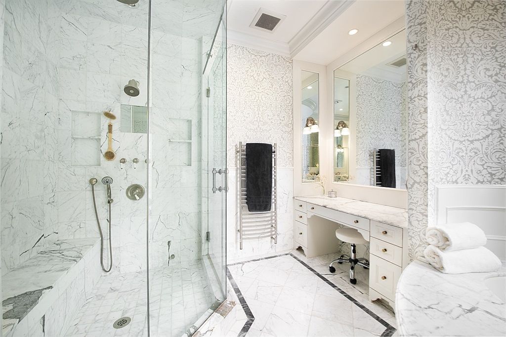 This luxurious primary bathroom features an expansive walk-in shower enclosed by glass, complete with a built-in stone bench and multiple shower heads. Adjacent to the shower is a dedicated vanity area with a stone countertop, a swivel stool, and a large mirror framed by elegant patterned wallpaper. The room is finished with light-toned stone flooring and a heated towel rack, creating a bright and sophisticated atmosphere.