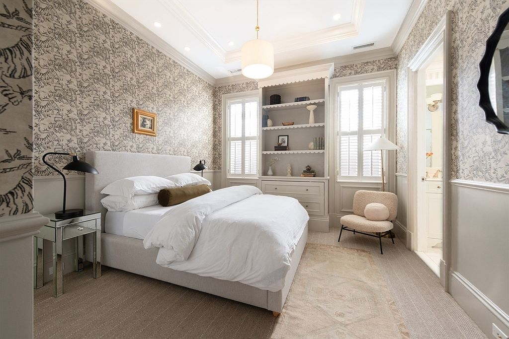 This elegantly appointed bedroom features a neutral color palette with sophisticated tiger-patterned wallpaper and a plush upholstered bed. A built-in shelving unit with a lower cabinet provides ample storage, flanked by two windows with white shutters that allow for soft, natural light. The room is completed by a mirrored nightstand, a textured area rug, and a cozy accent chair, creating a refined and tranquil atmosphere.