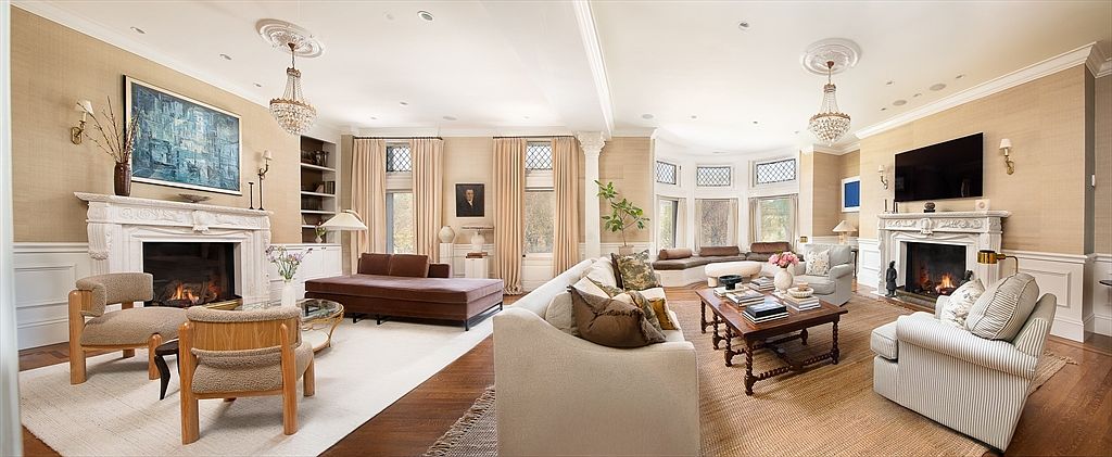 This expansive, light-filled living room features a sophisticated, traditional design with dual fireplaces and elegant chandeliers. The space is anchored by a large, neutral-toned sofa and a variety of seating arrangements, including a plush daybed and striped armchairs, all set against textured wall coverings. A wide bay window area with built-in bench seating provides a focal point, while the warm wood flooring and symmetrical layout create a balanced and inviting atmosphere.