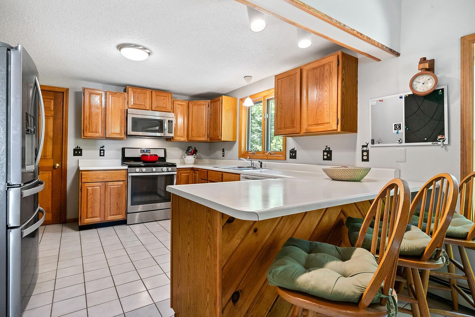 This kitchen features a functional layout with light wood cabinetry, a stainless steel refrigerator, and a matching microwave and range. A peninsula with a white countertop provides casual seating with wooden bar stools, while a white-tiled floor runs throughout the space. The room is illuminated by recessed lighting and a window above the sink, creating a bright and practical cooking environment.
