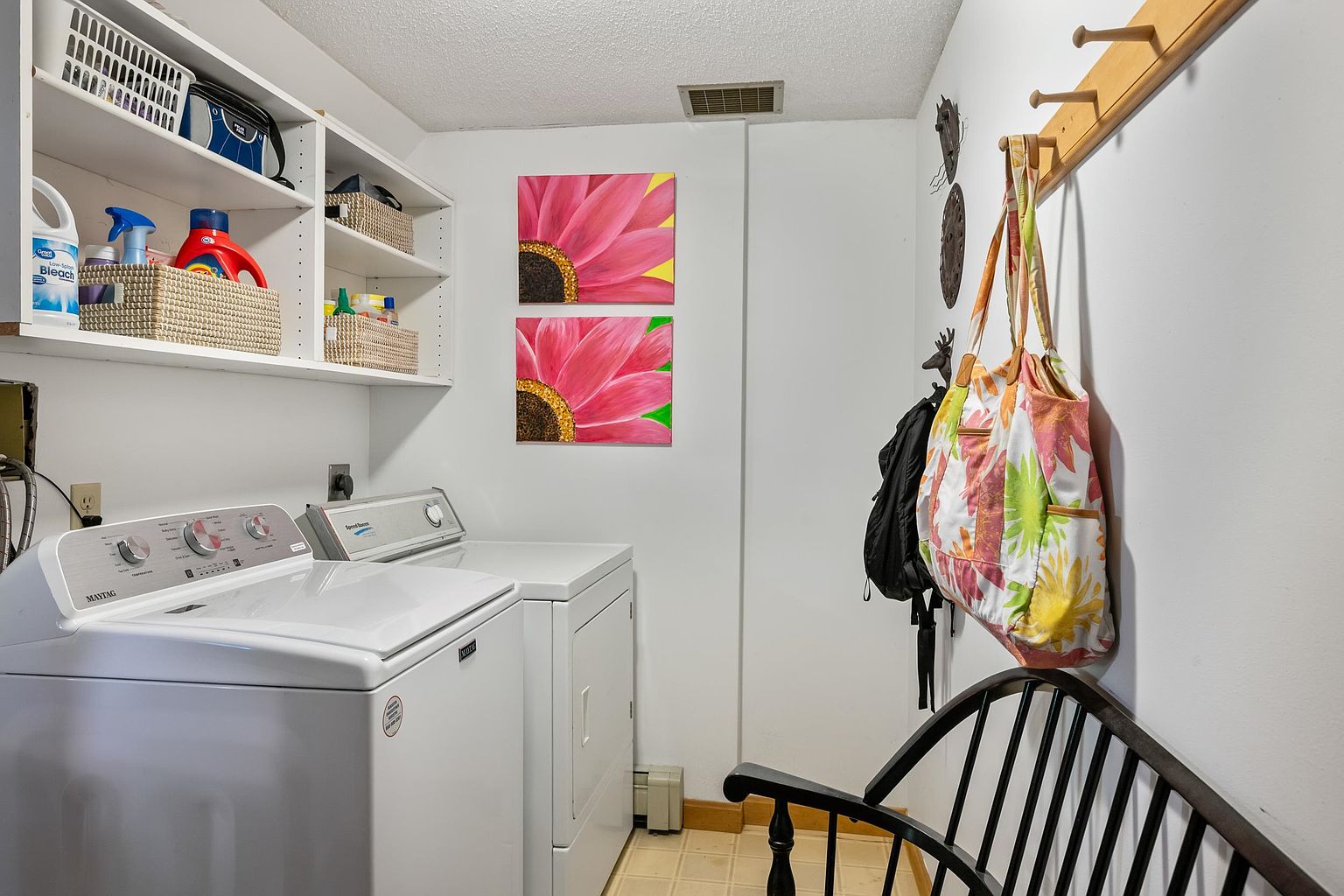 This functional laundry room features a white Maytag washing machine and a Speed Queen dryer positioned side-by-side. Above the appliances, white open shelving provides organized storage with woven baskets and cleaning supplies, while two vibrant floral canvas prints add a pop of color to the wall. A wooden coat rack with hanging bags and a dark wooden bench complete the space, creating a practical and tidy utility area.