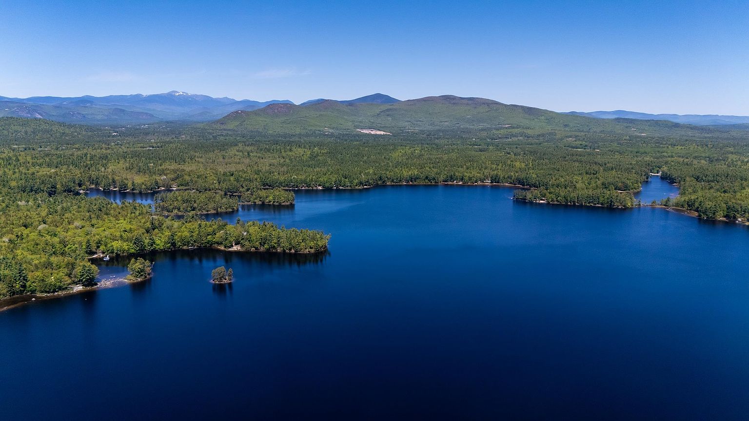 This high-altitude aerial perspective captures a serene, deep blue lake surrounded by a dense, lush green forest. In the distance, a range of rolling mountains under a clear sky provides a dramatic backdrop to the waterfront landscape. The composition emphasizes the expansive natural beauty and privacy of the location, highlighting the irregular shoreline and small islands dotting the water.