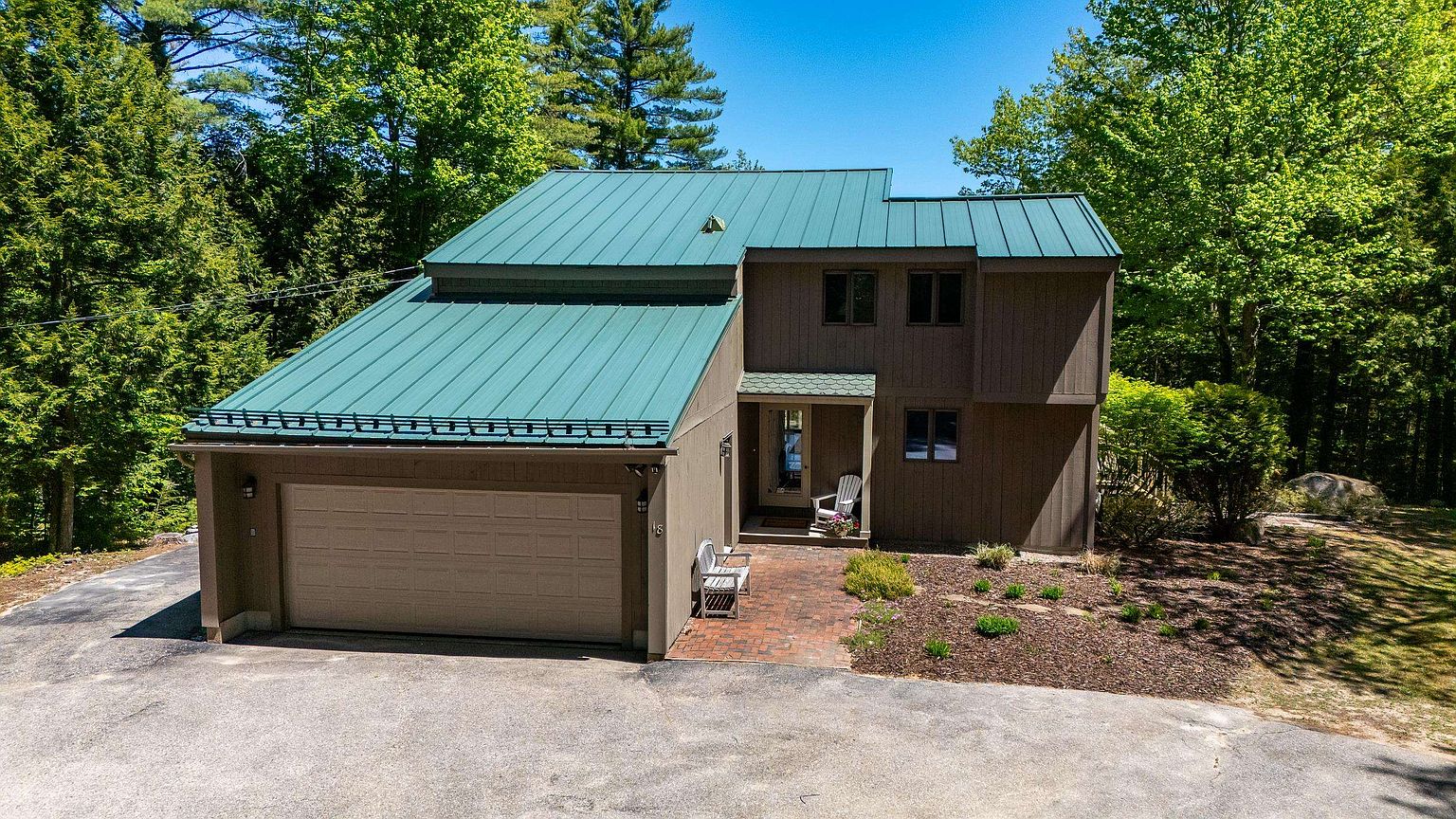This elevated, wide-angle shot captures the front facade of a two-story home nestled in a wooded setting. The house features a distinctive green metal roof, brown vertical siding, and an attached garage, with a brick-paved walkway leading to a covered entryway. The surrounding landscape includes mature trees and a natural, mulched garden bed, creating a serene and private atmosphere.