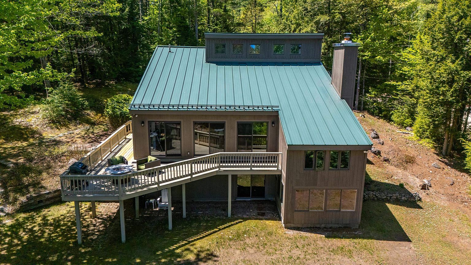 This aerial perspective captures a multi-level home nestled within a lush, wooded landscape. The structure features a prominent green metal roof, a large elevated deck with a fire pit, and a chimney stack rising along the side. The home's design incorporates a mix of vertical siding and large windows, creating a rustic yet modern aesthetic that blends seamlessly into the surrounding forest.