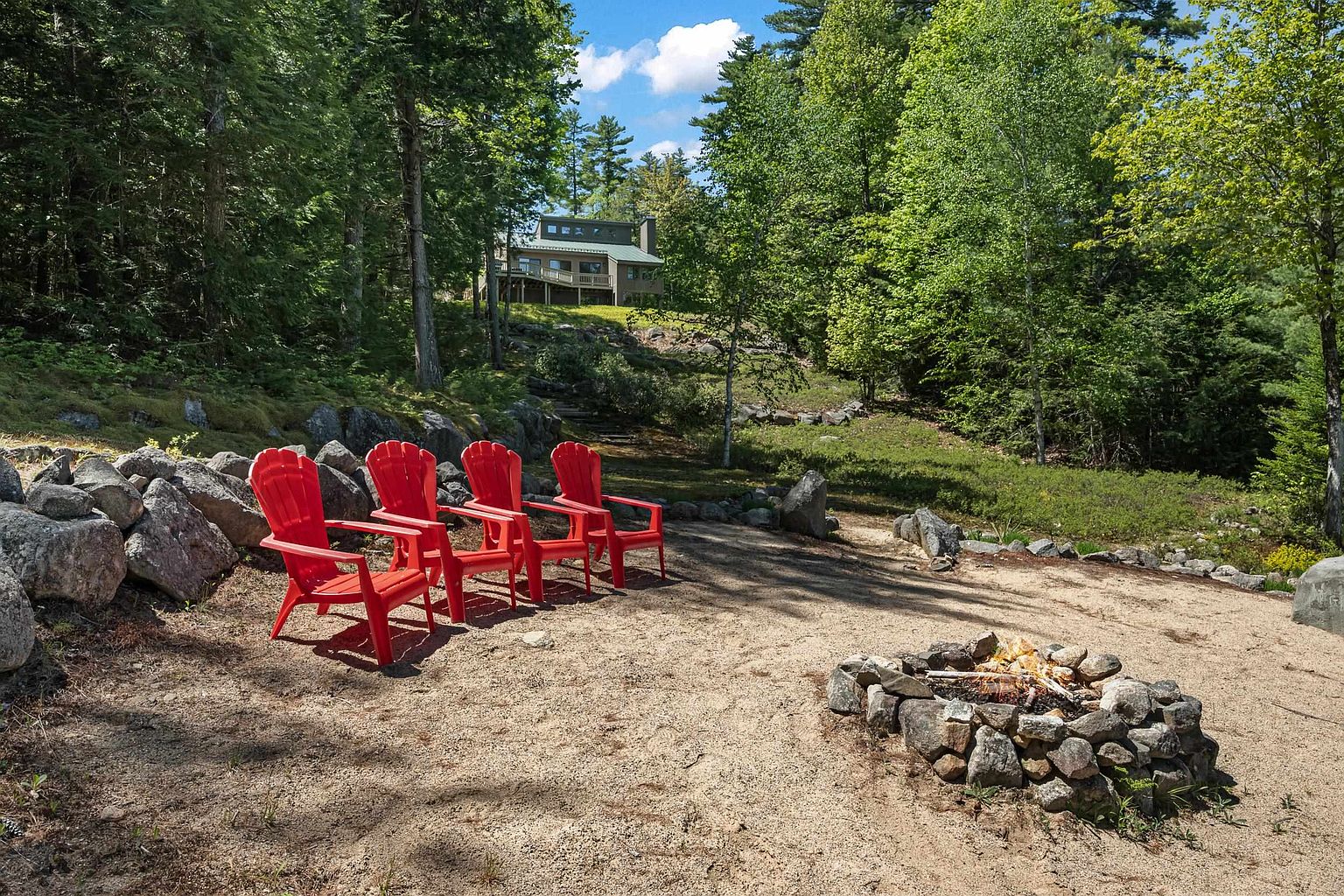 This scenic outdoor space features a sandy fire pit area equipped with four bright red Adirondack chairs, perfect for relaxing in a natural, wooded setting. A gentle slope leads up from the fire pit toward a multi-level house nestled among mature trees, creating a private and tranquil atmosphere. The landscape is accented by large, natural stone boulders and lush greenery, emphasizing the property's integration with the surrounding forest.