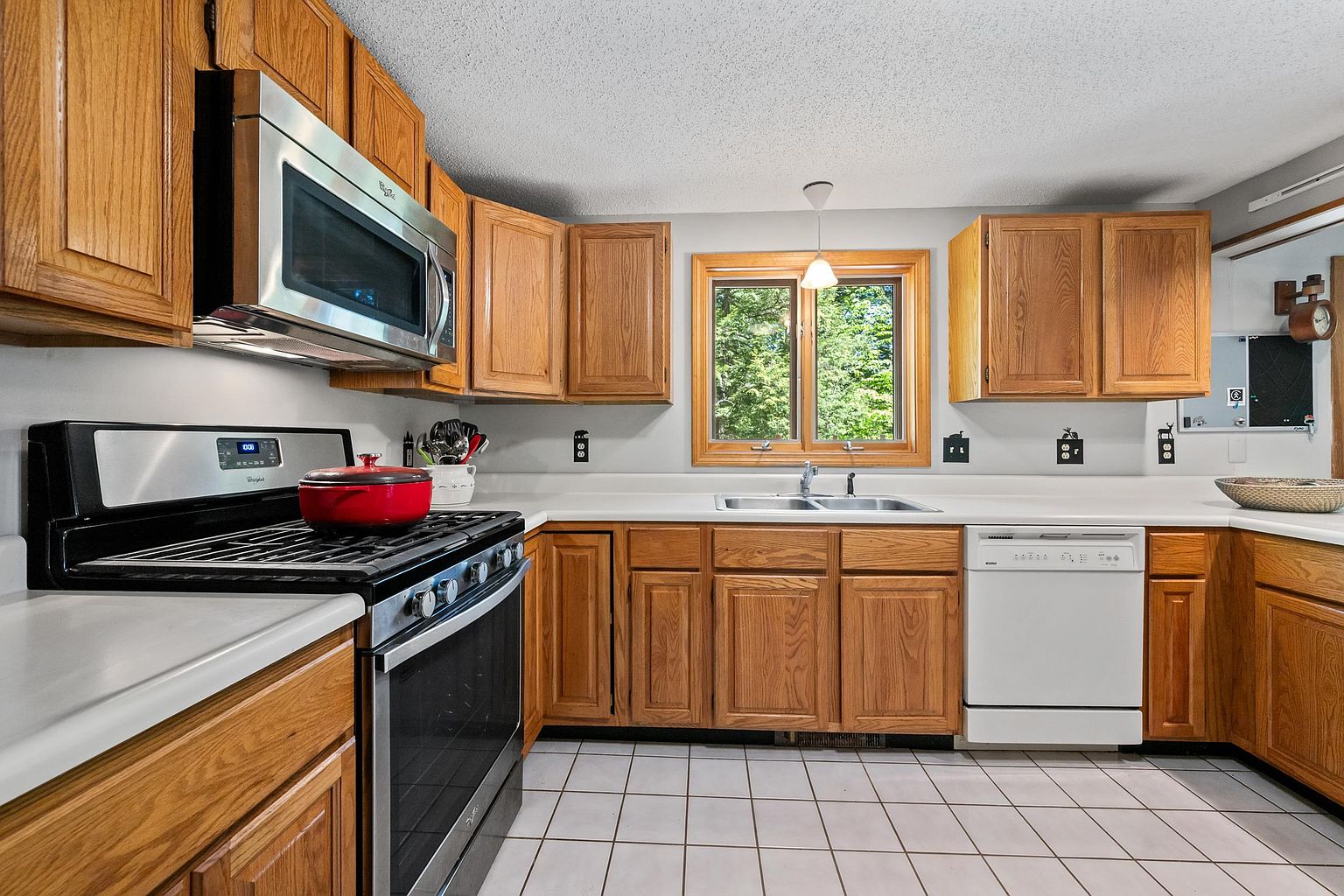This kitchen features traditional wood cabinetry, white countertops, and a suite of stainless steel appliances, including a gas range and a built-in microwave. A central window above the double-basin sink provides natural light, while the space is finished with light-colored floor tiles. The layout is functional and classic, offering a warm and inviting atmosphere for cooking and meal preparation.