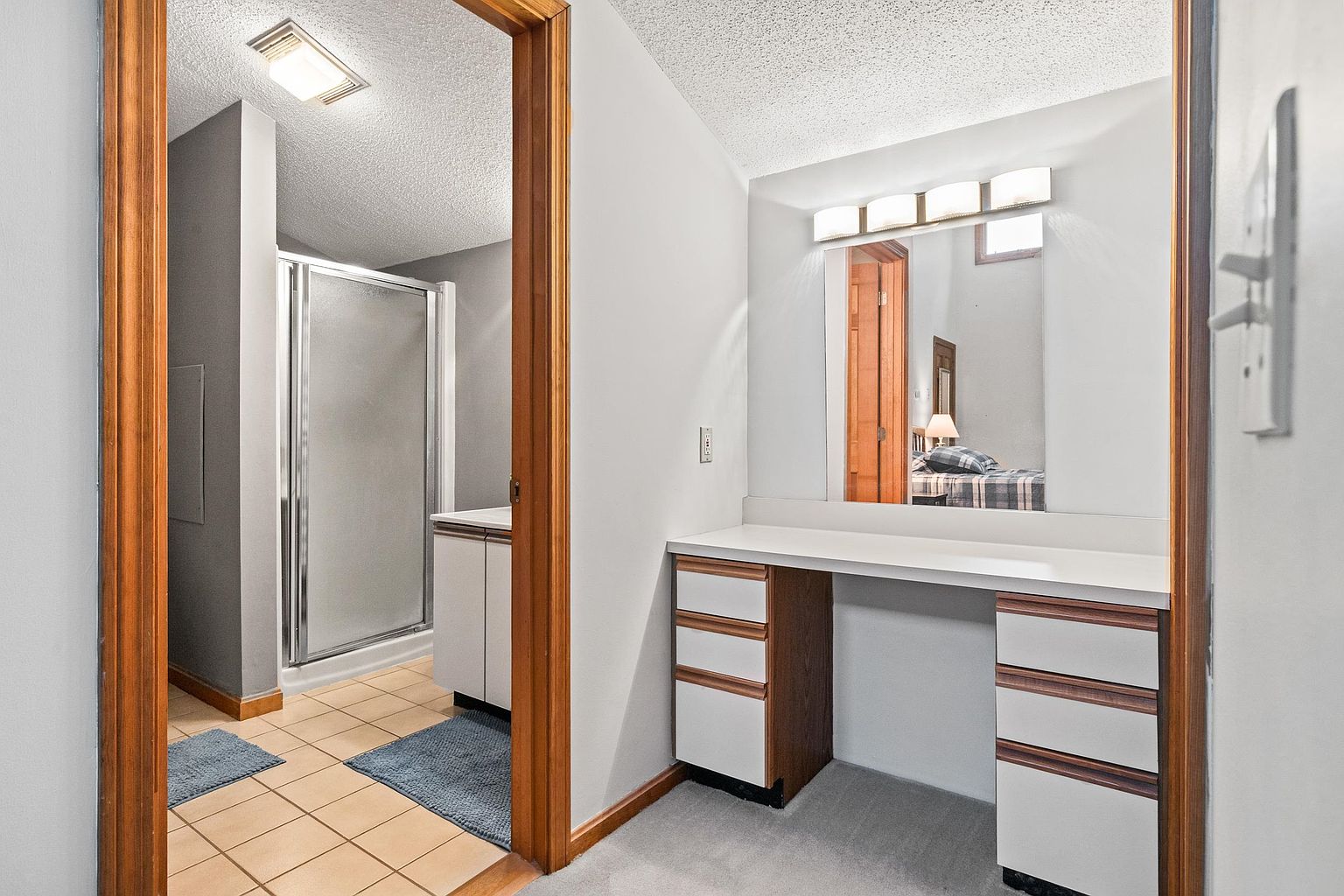 This primary bathroom suite features a vanity area with a white countertop and light-toned wood cabinetry, complemented by a large mirror and integrated lighting. A doorway leads into an adjacent tiled bathroom space containing a glass-enclosed shower and a secondary vanity. The neutral color palette and functional layout create a clean, practical, and well-organized space.
