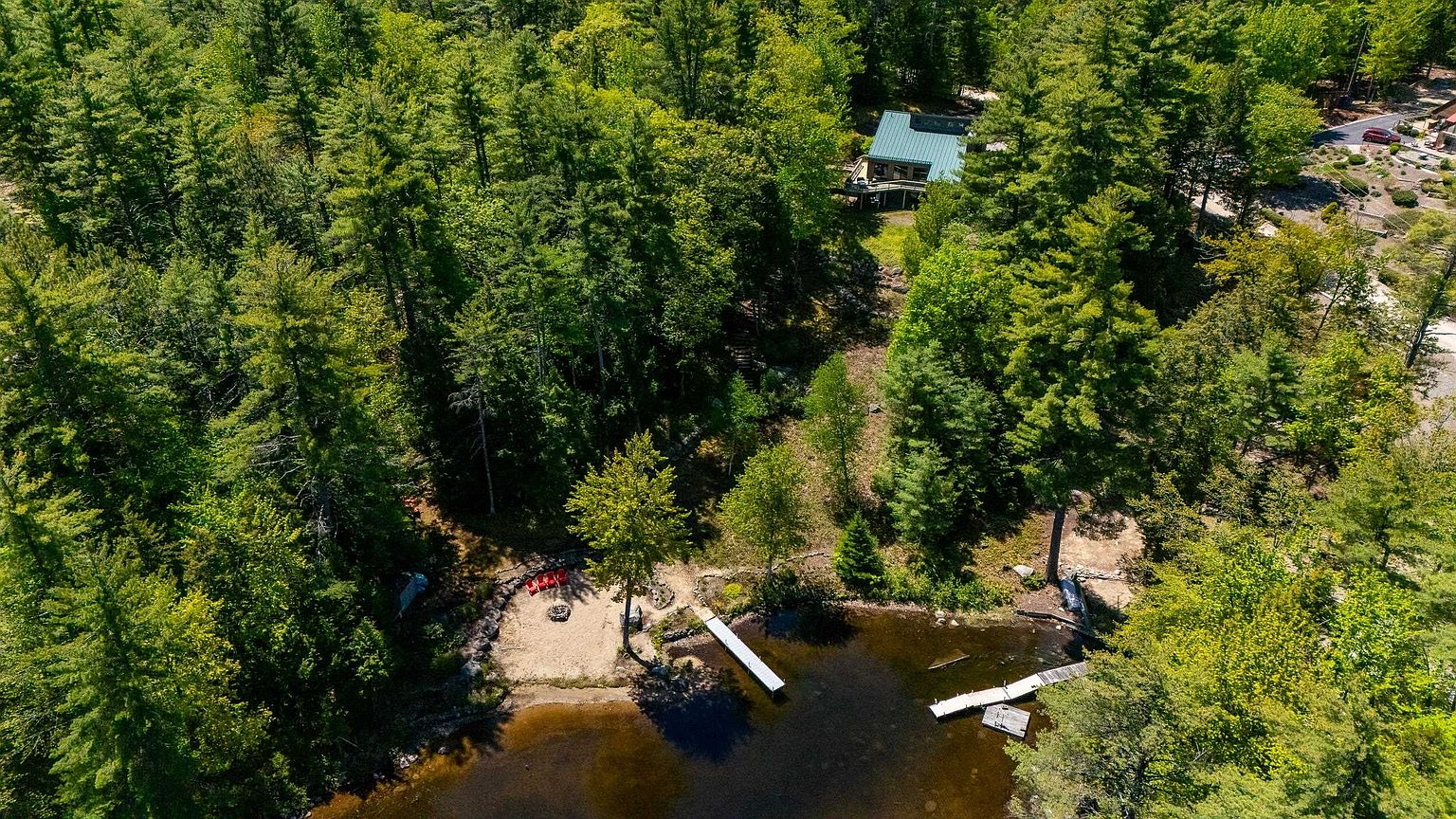 This high-angle aerial view captures a secluded waterfront property nestled within a dense, lush forest. The scene features a cabin with a green roof, a private sandy beach area with red chairs, and two wooden docks extending into the calm lake. The perspective emphasizes the property's peaceful, natural setting and direct access to the water.