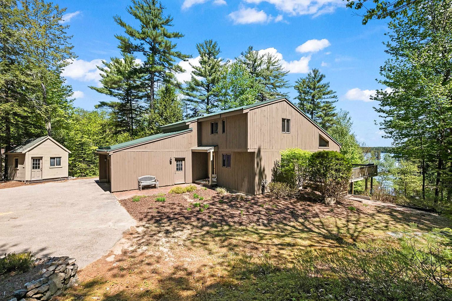 This side view of a multi-level home showcases its wood-sided exterior and a green metal roof, set against a backdrop of mature pine trees. A paved driveway leads to a detached shed, while a wooden deck extends from the side of the house toward a scenic lake view. The scene is captured in bright daylight, emphasizing the home's integration into its natural, wooded surroundings.