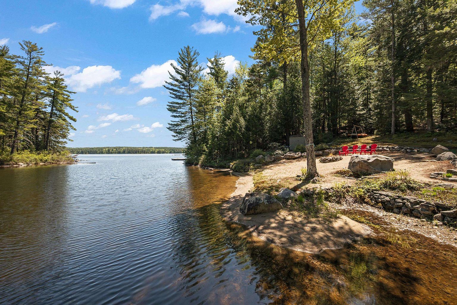 This serene waterfront property features a natural, sandy shoreline leading into a calm lake surrounded by dense, lush forest. A small clearing on the bank provides a peaceful outdoor seating area with several bright red chairs arranged near a large boulder. The perspective captures the tranquil transition from the water's edge to the wooded landscape, highlighting the property's private and scenic outdoor appeal.