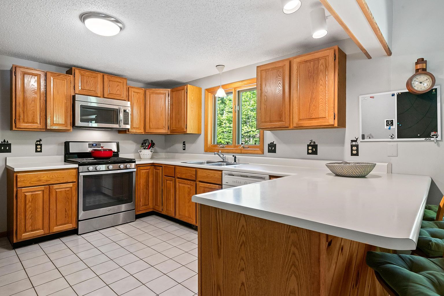 This kitchen features light wood cabinetry, white countertops, and a suite of stainless steel appliances, including a microwave and range. A peninsula provides additional counter space and seating, while a window above the sink allows natural light to brighten the room. The space is finished with light-colored floor tiles and a neutral wall color, creating a functional and traditional layout.