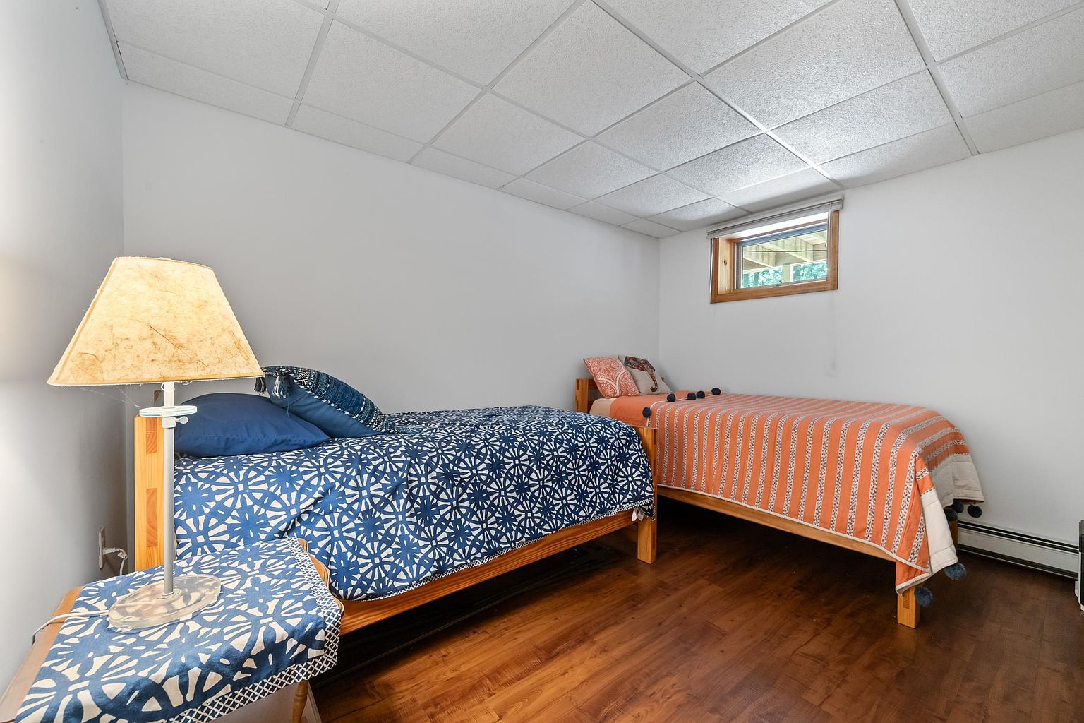 This bedroom features two twin-sized beds with wooden frames, one dressed in a blue patterned quilt and the other in an orange striped coverlet. The room has a drop-ceiling, white walls, and dark wood-look flooring, with a small window providing natural light. A bedside table with a lamp sits in the foreground, creating a cozy and functional space for guests.