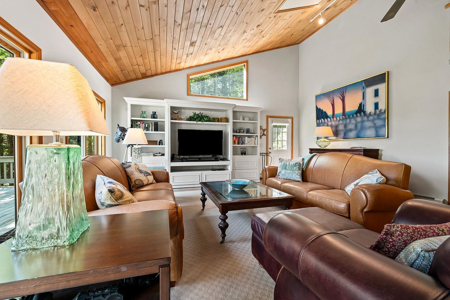This spacious living room features a vaulted wood-paneled ceiling and a large built-in media center with integrated shelving. The room is furnished with two tan leather sofas and a dark leather armchair, centered around a wooden coffee table with a glass top. A large, colorful landscape painting hangs on the wall, and the space is illuminated by multiple table lamps and natural light from a unique, angled window.
