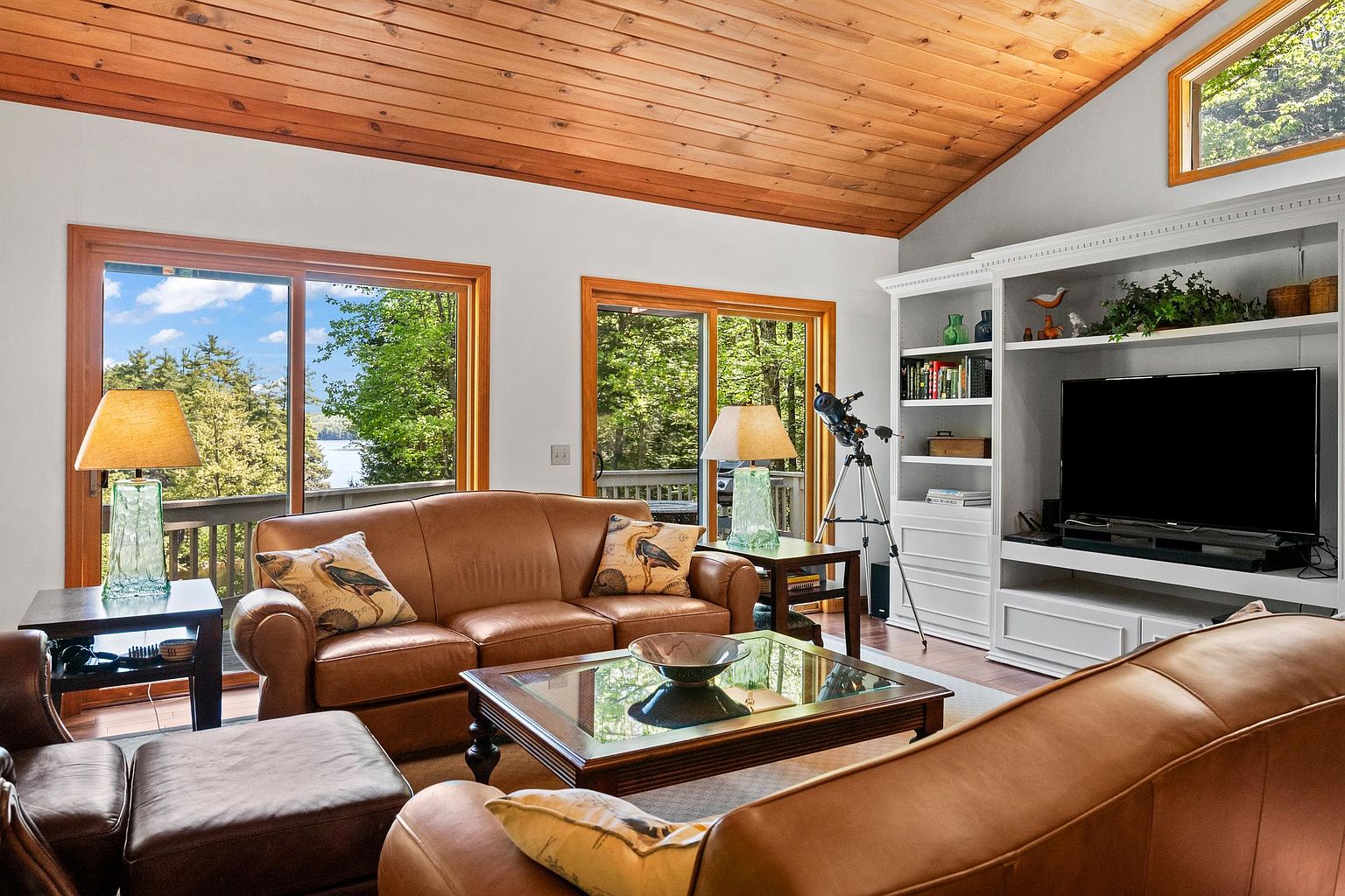 This inviting living room features a warm, wood-paneled vaulted ceiling and comfortable leather seating arranged around a glass-topped coffee table. Large sliding glass doors provide abundant natural light and scenic views of the surrounding trees and water. A built-in white shelving unit houses a television and decorative items, creating a cozy and functional space for relaxation.