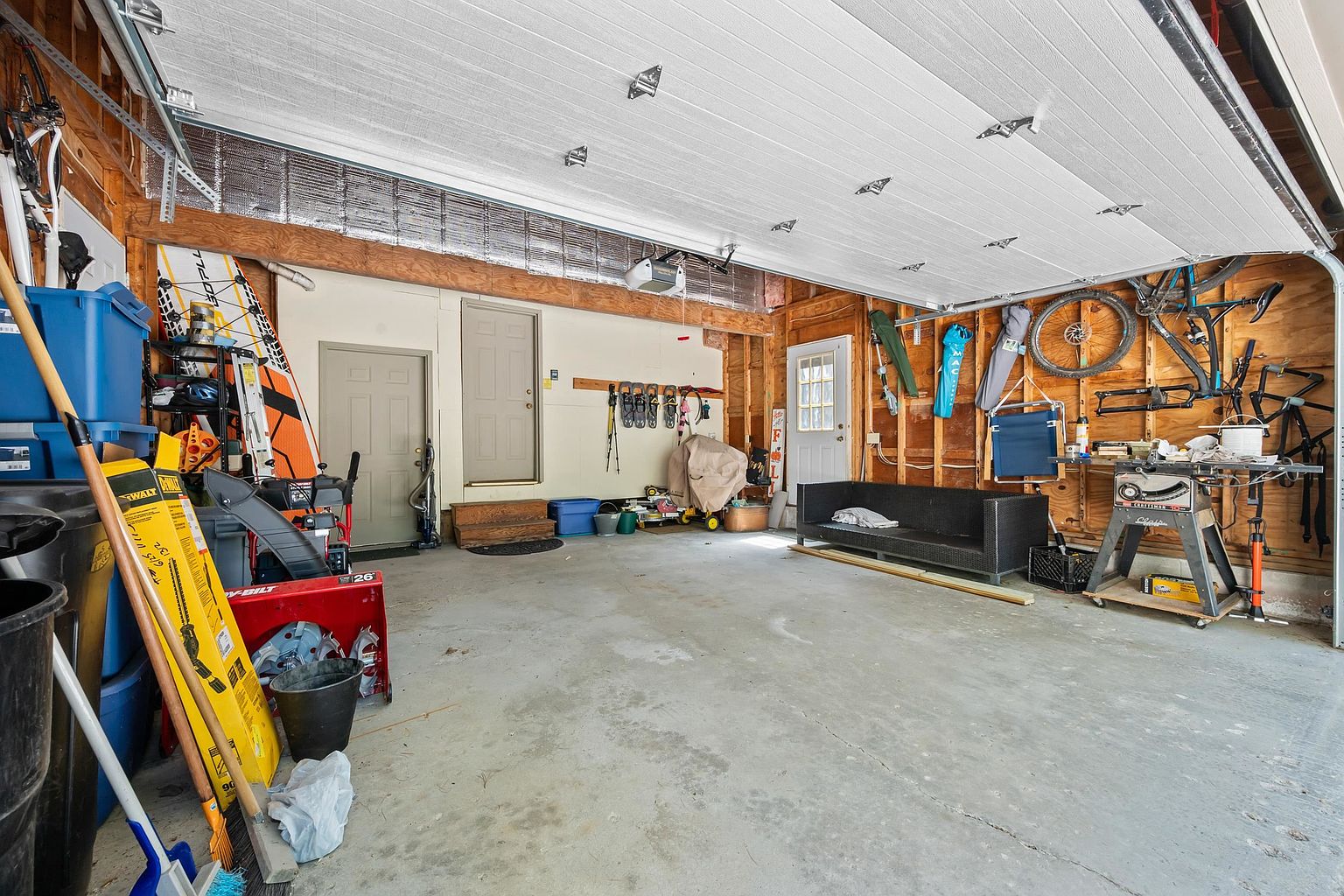 This spacious, unfinished garage features exposed wood framing and a concrete floor, offering ample room for storage and projects. The space is equipped with a workbench, wall-mounted bicycle storage, and multiple entry doors leading to other parts of the home. A black sofa sits against the wall, providing a casual seating area within this functional utility space.