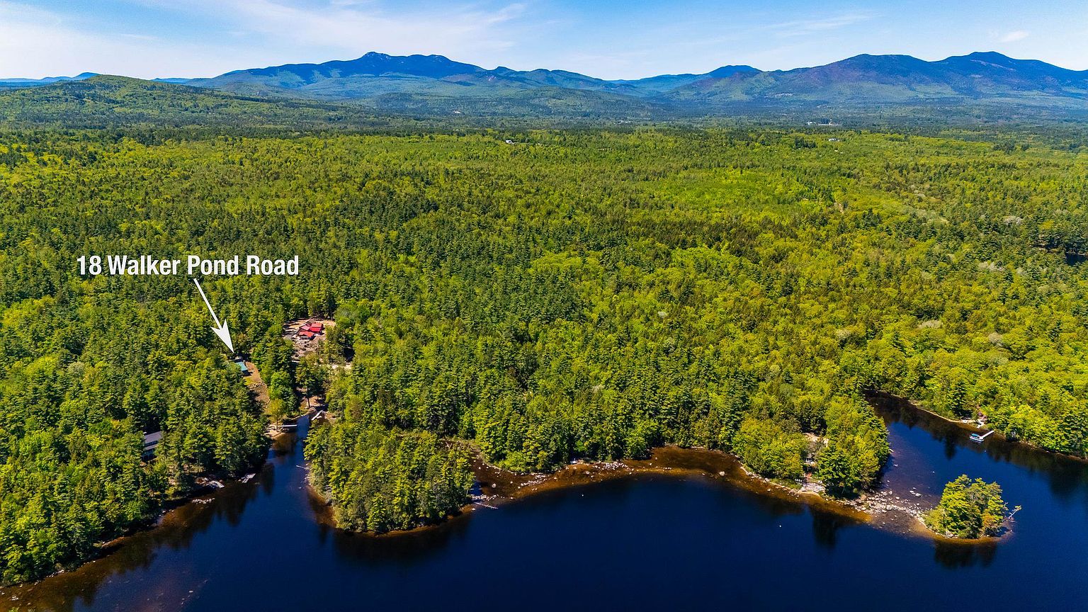 This high-altitude aerial view captures a secluded property nestled within a dense, lush forest bordering a serene pond. The landscape features rolling hills and distant mountains in the background, emphasizing the property's private and natural setting. A small structure is visible among the trees, highlighting the remote and tranquil nature of the location.