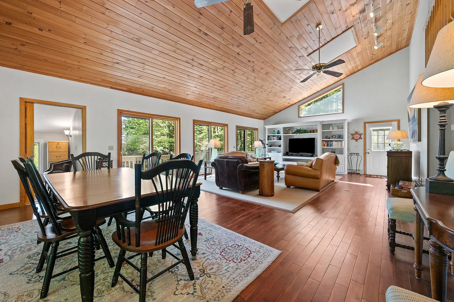 This open-concept living and dining area features a dramatic vaulted ceiling with exposed wood paneling and a ceiling fan. The space is anchored by a dining set in the foreground and a comfortable seating area with leather furniture arranged around a media center in the background. Large sliding glass doors provide ample natural light and views of the surrounding greenery, creating a warm and inviting atmosphere.