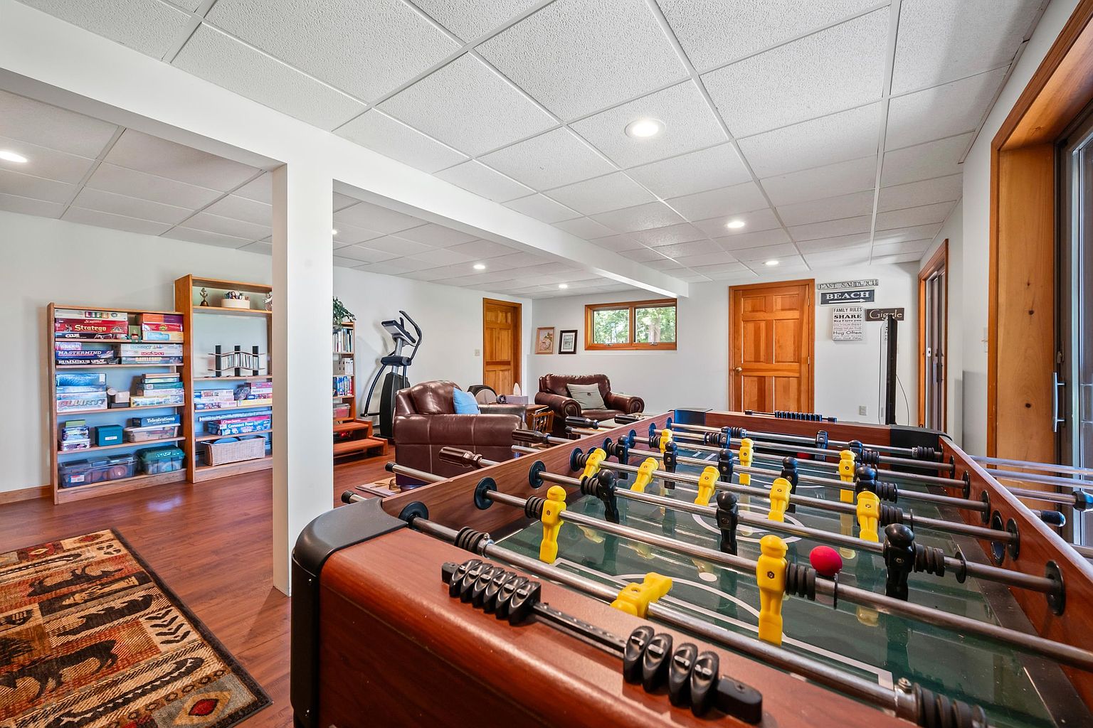 This spacious finished basement features a dedicated game area with a foosball table in the foreground and a comfortable seating area with leather armchairs in the background. The room includes built-in shelving units filled with board games, wood-look flooring, and a drop ceiling with recessed lighting. A sliding glass door provides natural light, creating an inviting and functional recreational space.