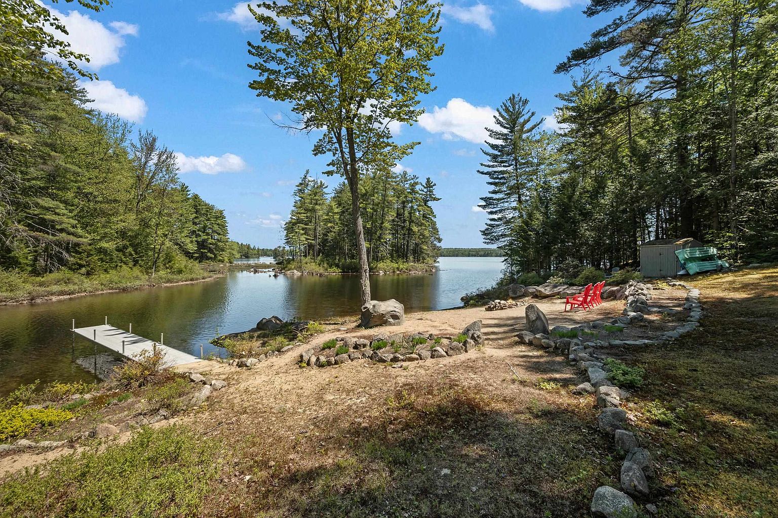 This scenic waterfront property features a natural, sandy shoreline leading directly to a calm lake with a private wooden dock. The landscape is framed by lush, mature trees and includes a stone-lined path, a fire pit area with bright red Adirondack chairs, and a small storage shed. The perspective captures the serene, expansive outdoor living space, highlighting the peaceful connection between the wooded lot and the water.