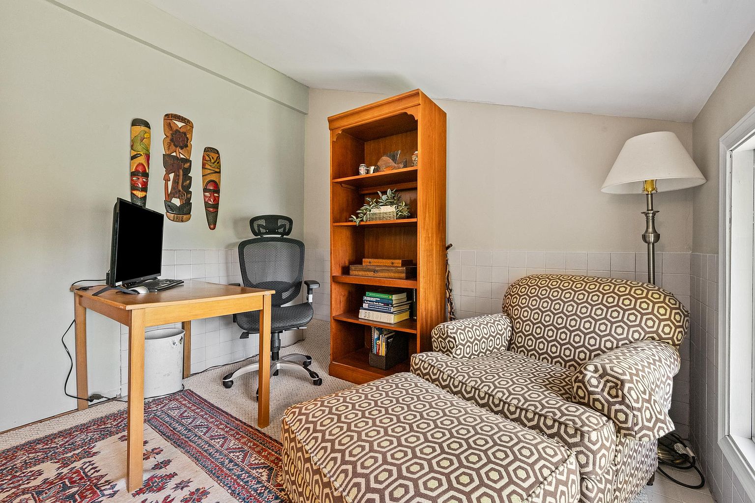 This cozy home office space features a light wood desk paired with an ergonomic mesh chair, positioned near a wall decorated with three vertical wooden art pieces. A tall, light wood bookshelf provides storage for books and decor, while a comfortable armchair with a matching ottoman sits in the corner next to a floor lamp. The room is grounded by a patterned area rug, creating a functional and inviting workspace.