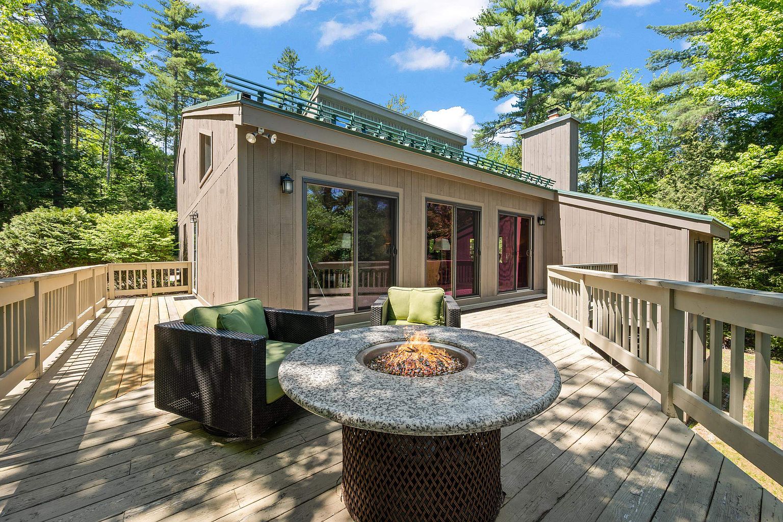 This spacious wooden deck features a central circular stone fire pit with a glowing flame, surrounded by two dark wicker armchairs with green cushions. The deck is enclosed by wooden railings and provides a clear view of the home's exterior, which includes large sliding glass doors and a tall chimney. The setting is nestled among lush green trees, creating a serene and private outdoor living space perfect for relaxation.
