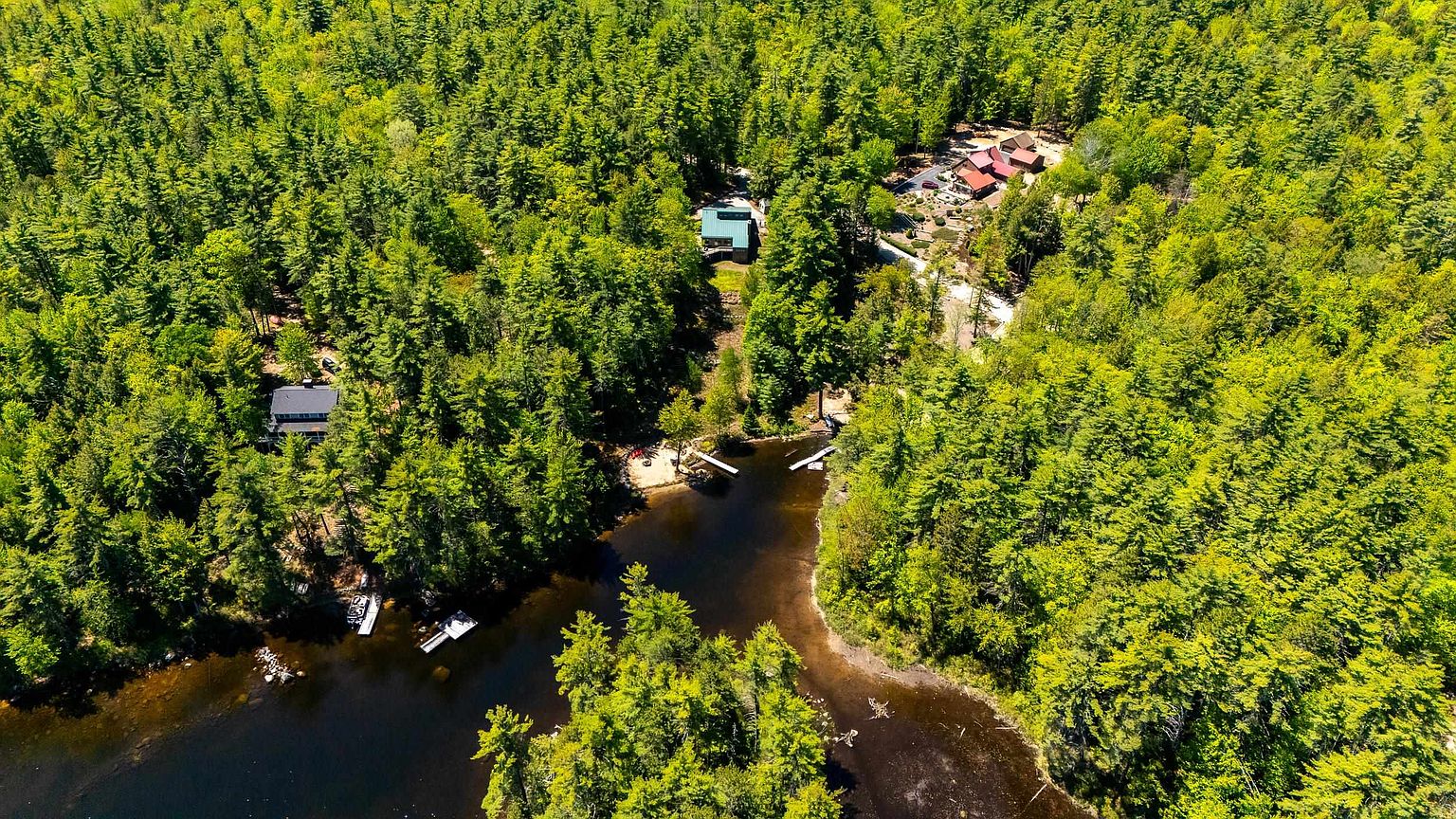 This high-angle aerial view captures a secluded woodland property nestled within a dense forest. The scene features multiple structures, including a modern home and a larger residential complex, situated near a tranquil, dark-water lake with a private dock and sandy shoreline. The lush green canopy provides a sense of privacy and natural beauty, highlighting the property's serene, waterfront lifestyle.