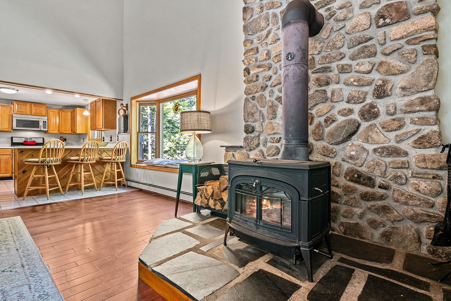 This inviting living space features a prominent wood-burning stove set against a rustic stone-clad chimney breast, resting on a raised slate hearth. To the left, an open-concept kitchen with light wood cabinetry and a breakfast bar with three stools overlooks the living area, which is illuminated by a large bay window with a built-in bench seat. The room is finished with warm-toned wood flooring, creating a cozy and cohesive atmosphere that blends rustic charm with functional living space.