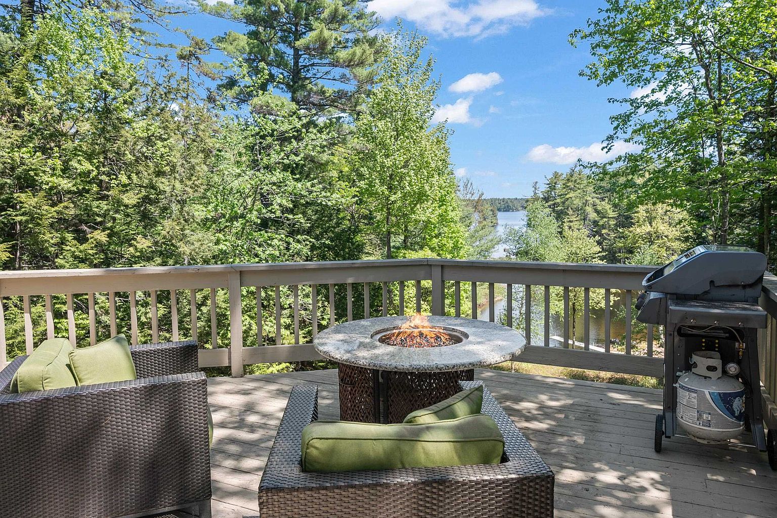This elevated wooden deck offers a serene outdoor living space featuring a circular stone-topped fire pit surrounded by comfortable wicker armchairs with green cushions. To the right, a gas grill is positioned near the railing, which overlooks a lush, tree-lined landscape and a glimpse of a lake in the distance. The perspective is from a slightly elevated eye-level, capturing the inviting atmosphere of a private retreat perfect for relaxing or entertaining.