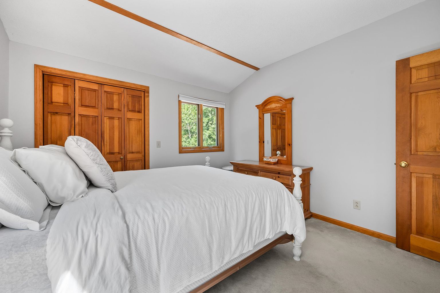 This bedroom features a neutral color palette with light gray walls and a vaulted ceiling accented by a wood beam. The room is furnished with a bed dressed in white linens and a wooden dresser topped with a mirror, complemented by matching wooden closet doors and a door leading to the rest of the home. A window centered on the wall provides natural light, creating a bright and comfortable atmosphere.