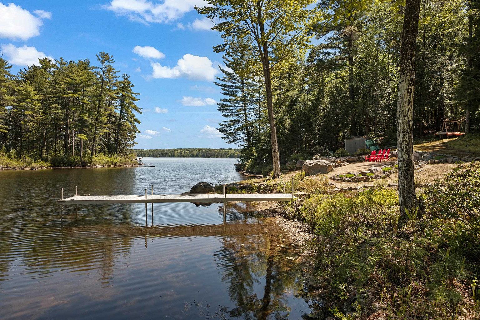 This serene waterfront scene features a long, narrow dock extending into a calm, clear lake surrounded by dense, lush forest. The shoreline includes a natural, rustic landscape with large boulders, native shrubs, and a small clearing furnished with bright red Adirondack chairs. The perspective captures the peaceful transition from the wooded property to the open water under a bright, partly cloudy sky.
