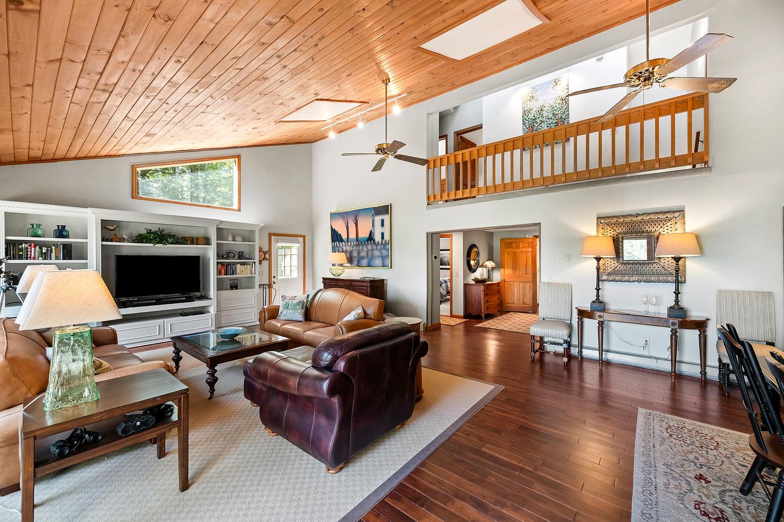 This spacious, open-concept living room features a dramatic vaulted wood-paneled ceiling and a mezzanine balcony overlooking the main floor. The room is furnished with comfortable leather seating, a central coffee table, and built-in shelving surrounding a television, creating a warm and inviting atmosphere. A wide-angle perspective captures the flow between the living area, the dining space, and the hallway leading to other parts of the home.