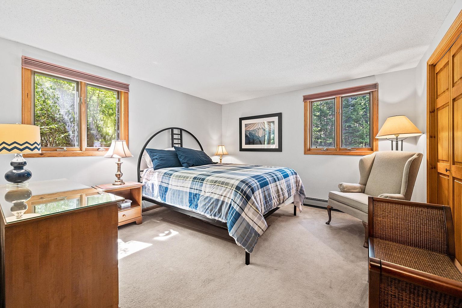 This cozy bedroom features a metal-framed bed dressed in a blue and white plaid comforter, positioned centrally between two windows that offer views of the surrounding greenery. The room includes a light-colored wingback chair, a wooden dresser with a glass top, and a matching nightstand, all set against neutral walls and carpeted flooring. The space is illuminated by multiple lamps, creating a warm and inviting atmosphere suitable for a guest suite.